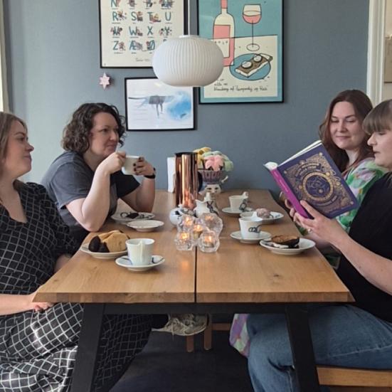 Fire bibliotekarer sidder ved et bord og drikker kaffe, mens den ene læser højt af en bog