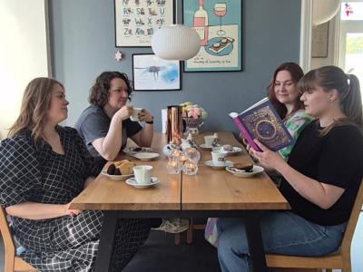 Fire bibliotekarer sidder ved et bord og drikker kaffe, mens den ene læser højt af en bog