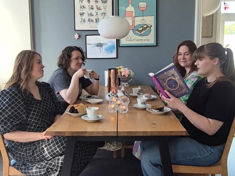 Fire bibliotekarer sidder ved et bord og drikker kaffe, mens den ene læser højt af en bog